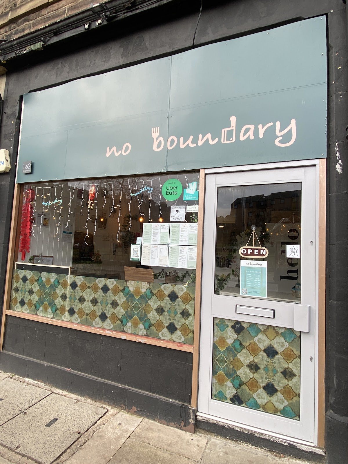 The shopfront on Gorgie Road: a teal fascia with 'no boundary' in pink lowercase, a window of blue-green patterned tiles below, and a white door with an OPEN sign and 'hello' on the glass.