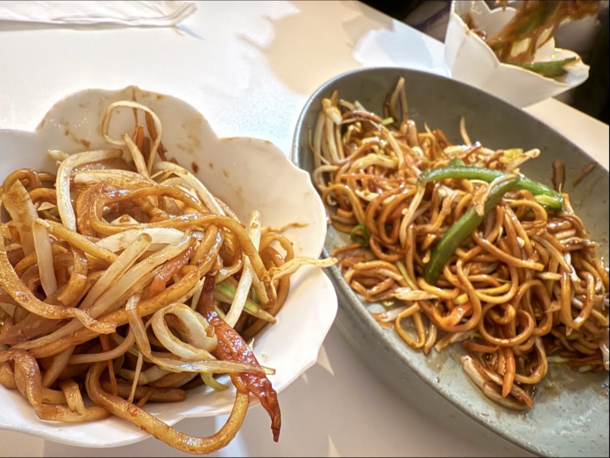 Two plates of glossy wok-fried noodles, one with beansprouts and spring onion, one with strips of green pepper.